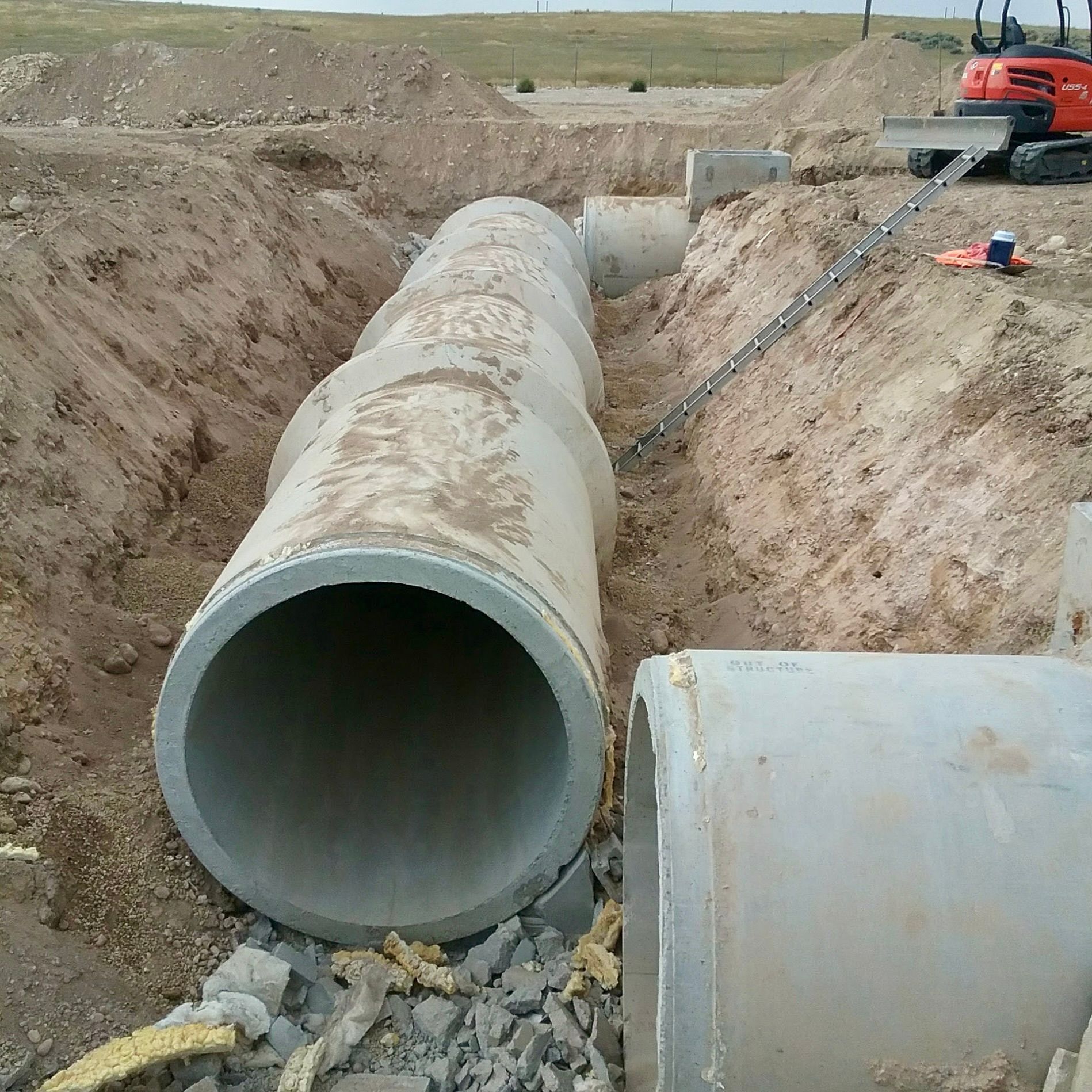 Large concrete pipes in a trench at a construction site. An excavator and ladder are nearby.