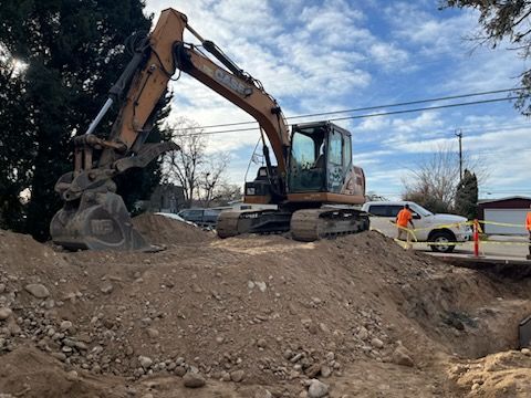 An excavator digs into dirt pile, near white truck and workers in orange vests, under a cloudy sky.