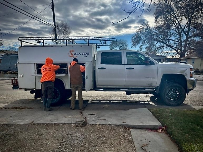 Two people near a white work truck with side storage. One in orange, one in brown, both near open doors. Cloudy sky.