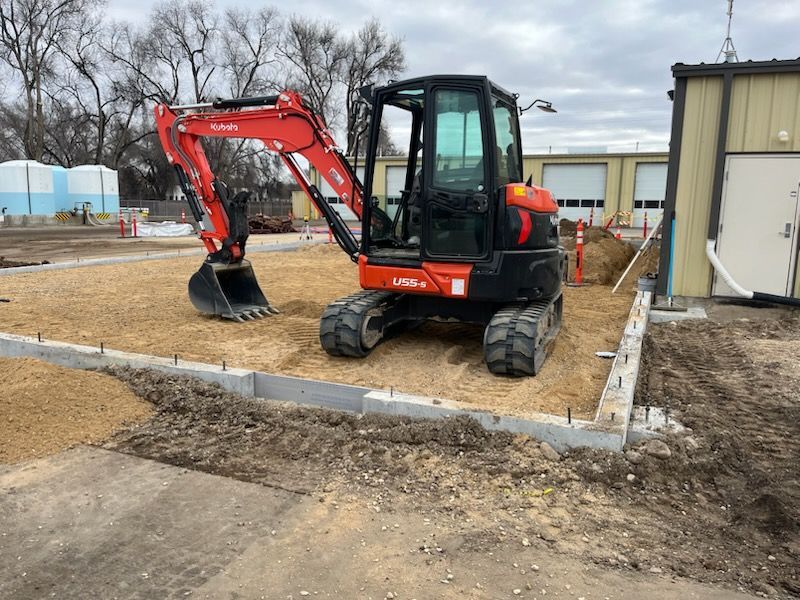 Mini excavator on gravel within a concrete frame, near a building.