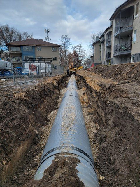 Large gray pipe in a deep trench at a construction site near apartment buildings.