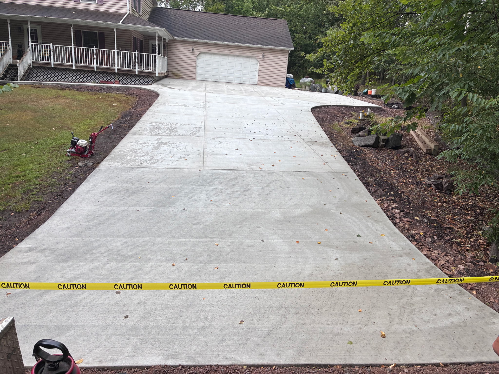 Concrete driveway with visible cracks, sloping up to a house garage, bordered by landscaping and caution tape at the bottom.