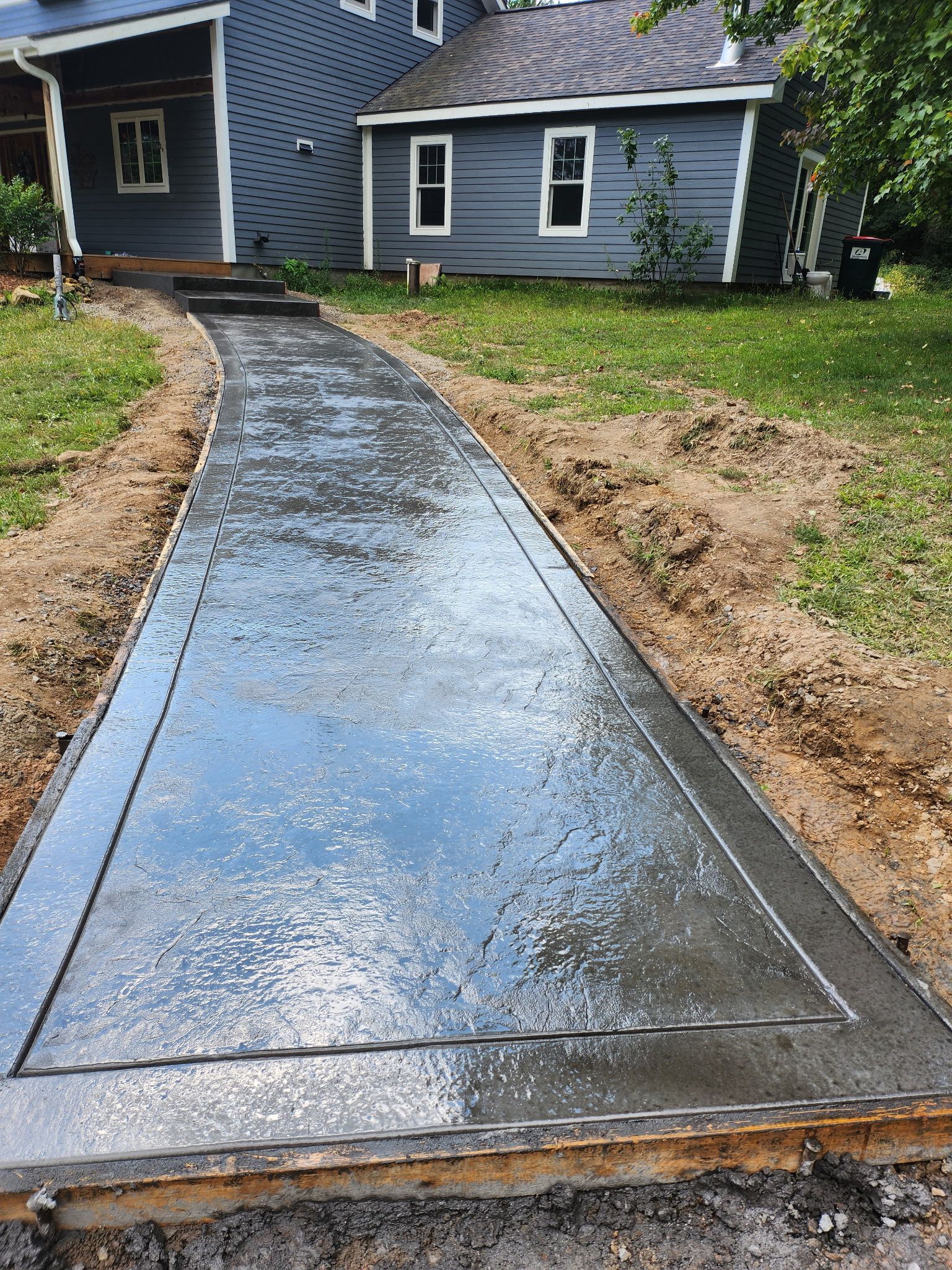 Wet concrete walkway leading to a blue house with grass on both sides