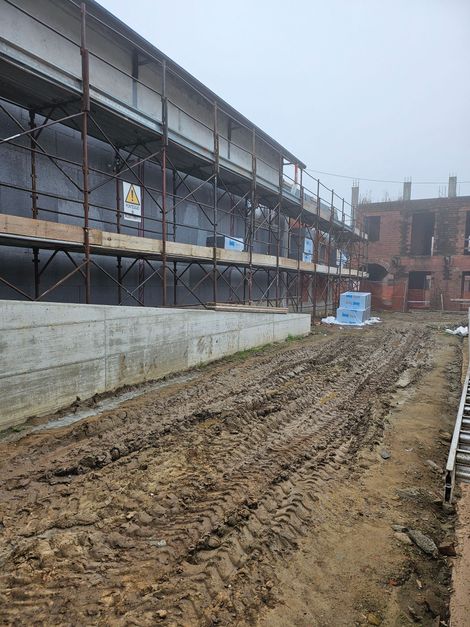 Cantiere edile ricoperto di fango con impalcature lungo un edificio e un sentiero di terra battuta bagnato.