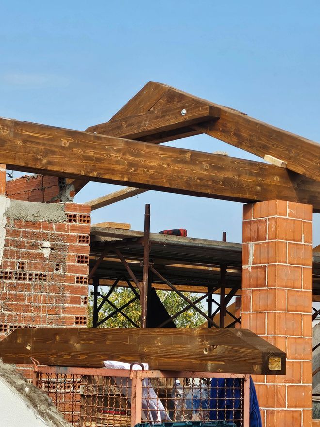 Struttura in mattoni parzialmente costruita con travi del tetto in legno, stagliata contro un cielo limpido.