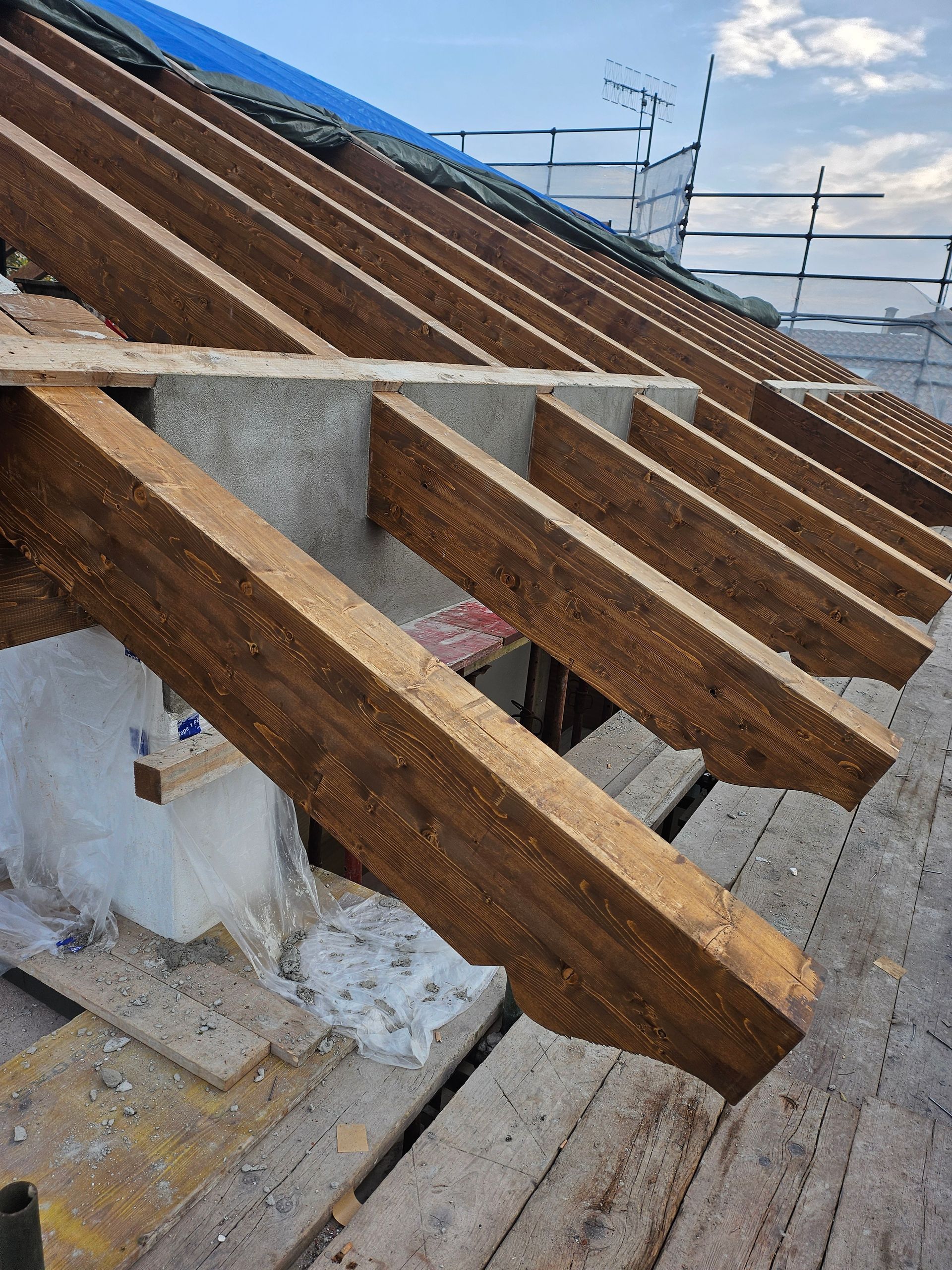 Travi di legno sul tetto in fase di costruzione, con supporti in cemento e impalcature degli operai sullo sfondo.