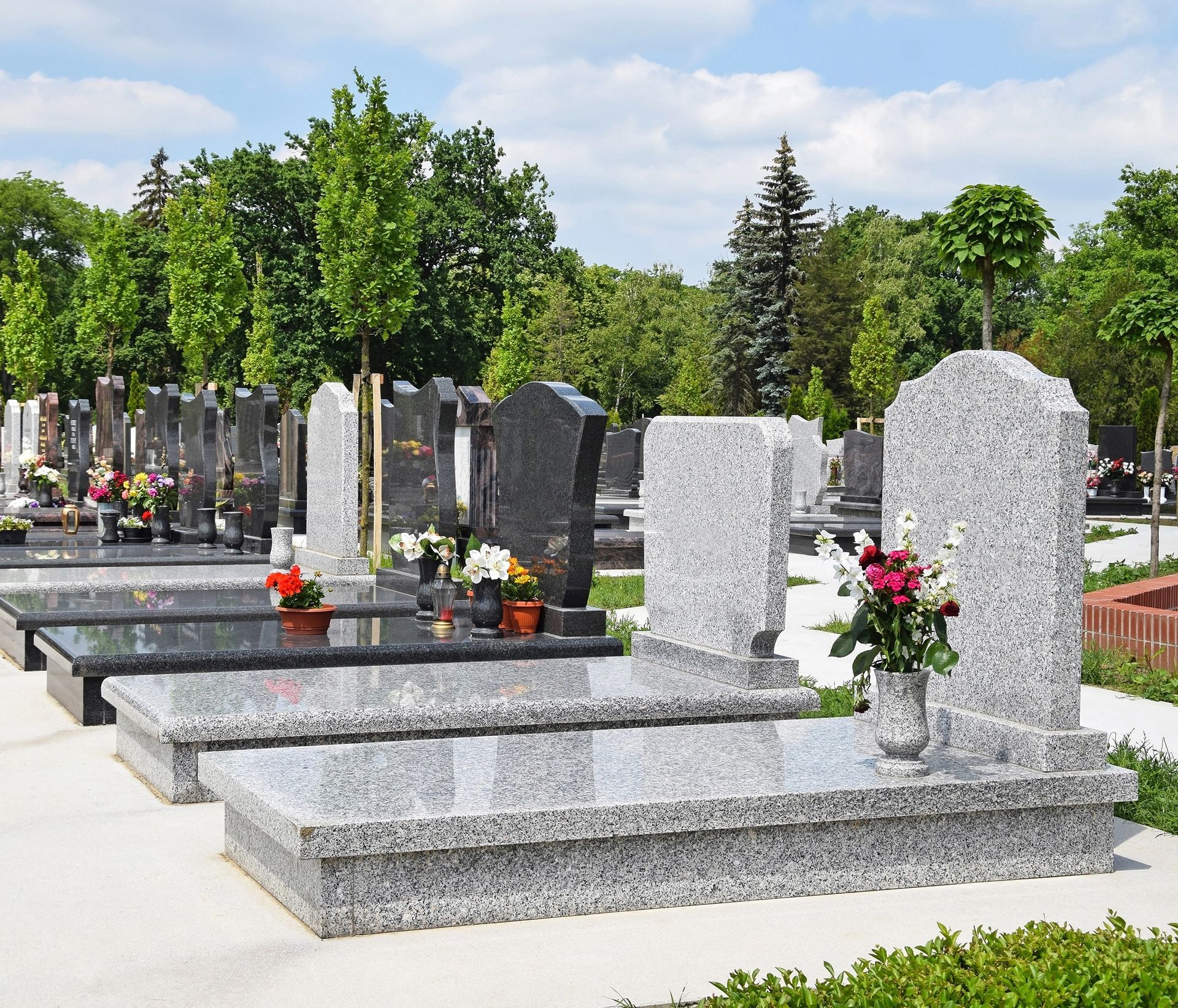 File di lapidi grigie con vasi di fiori in un cimitero soleggiato