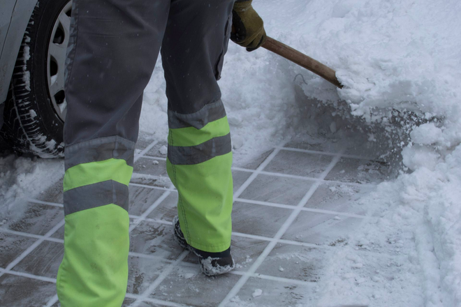 Una persona sta spalando la neve da un marciapiede, con pantaloni verde brillante e neve accumulata nelle vicinanze.