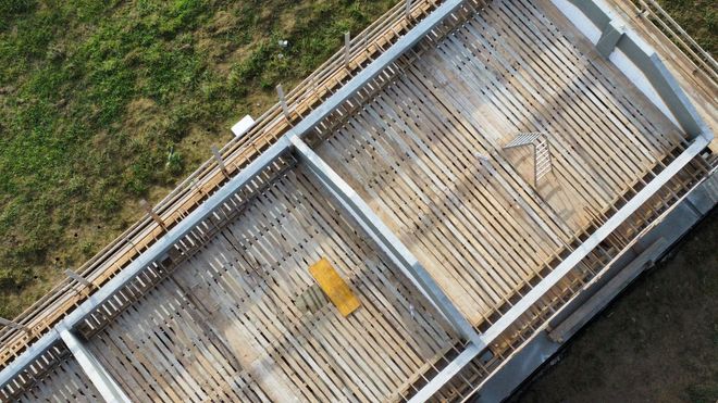 Veduta aerea di un ponte di legno consumato dal tempo che attraversa un prato, con un piccolo indicatore giallo al centro.