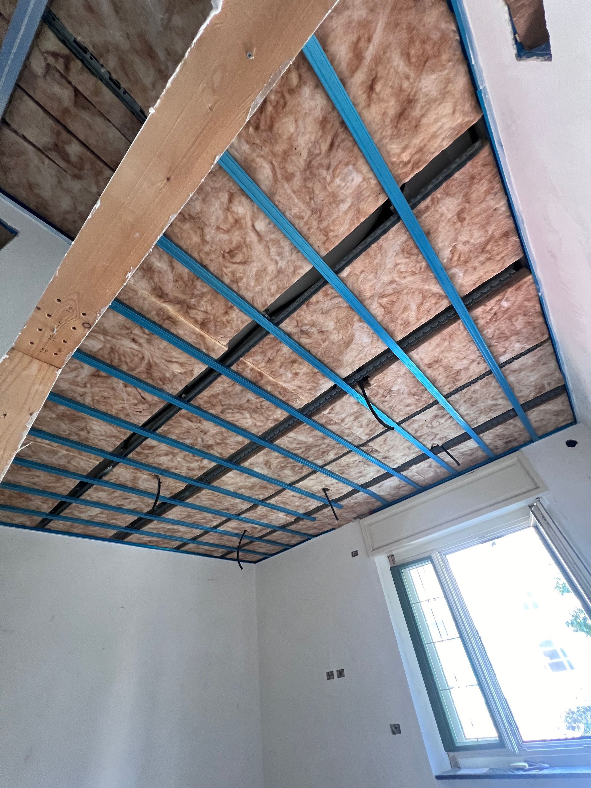 Struttura del soffitto a vista con isolamento rosa e travetti blu in una stanza con pareti bianche e una finestra.