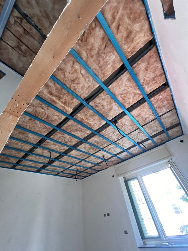 Struttura del soffitto a vista con isolamento rosa e travetti blu in una stanza con pareti bianche e una finestra.