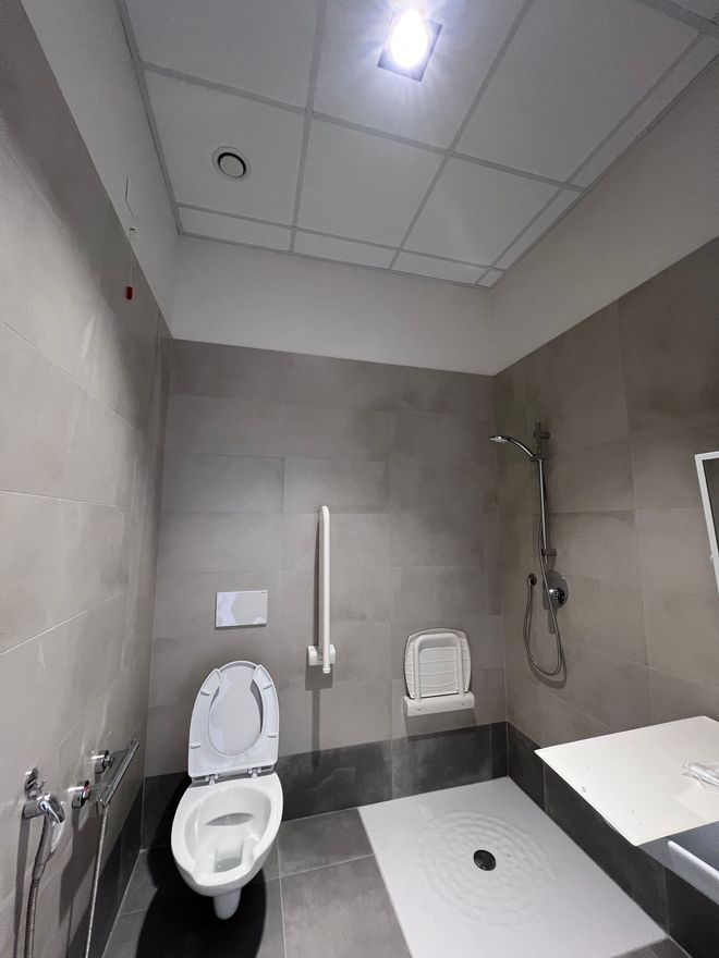 Piccolo bagno accessibile con piastrelle grigie, dotato di WC, maniglioni di sostegno, lavandino e sedile doccia a parete.