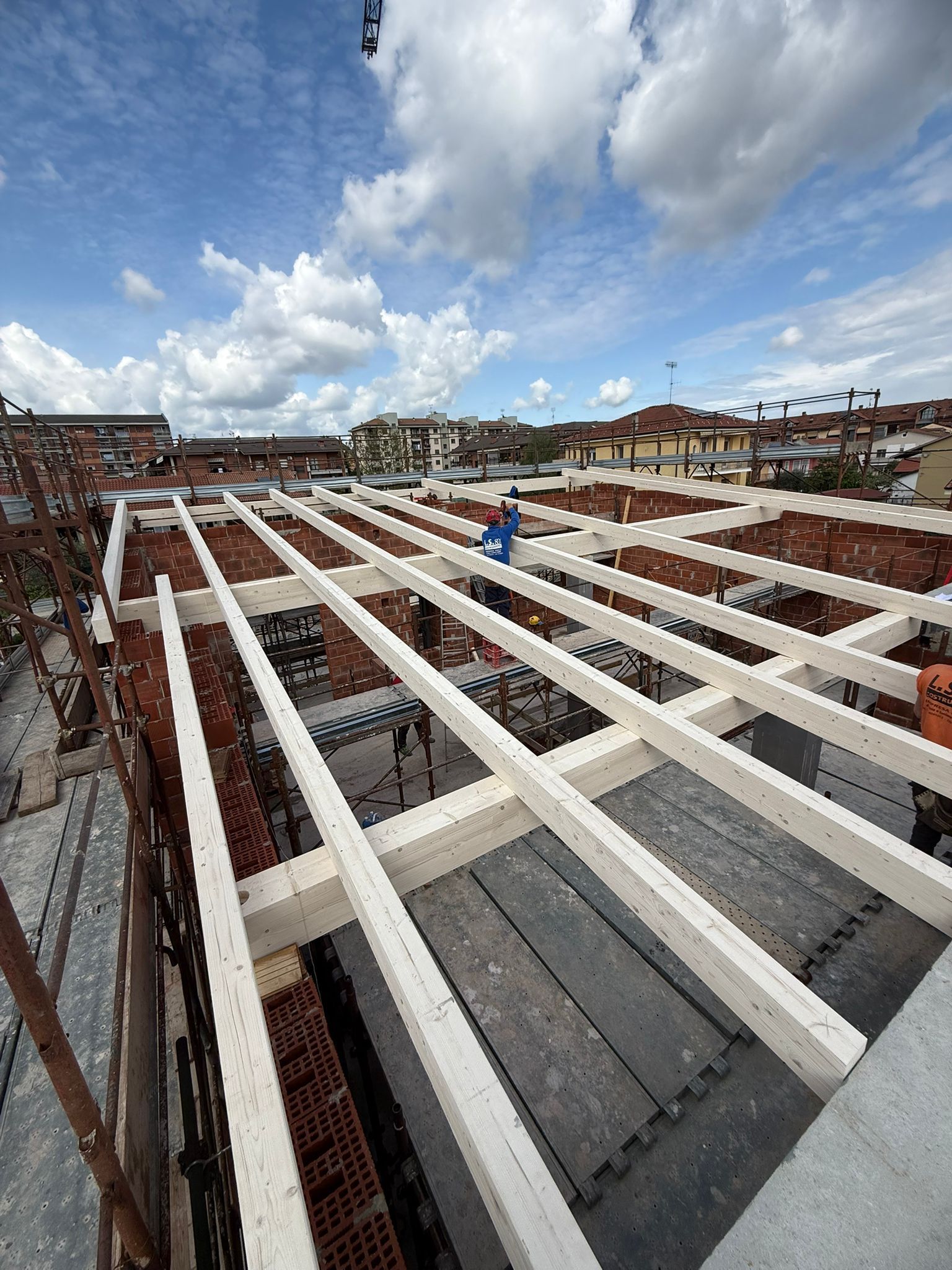 Cantiere edile con travi bianche sul tetto di una struttura in mattoni sotto un cielo nuvoloso.