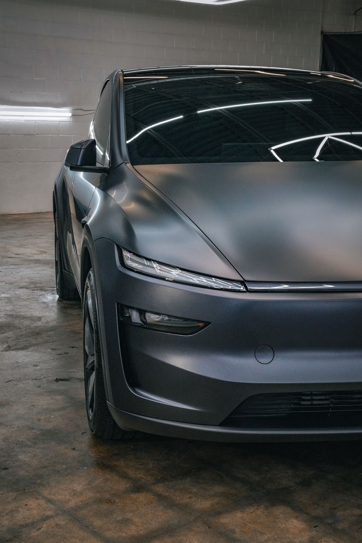 A matte black Tesla Model Y parked inside a workshop with reflective overhead lighting.