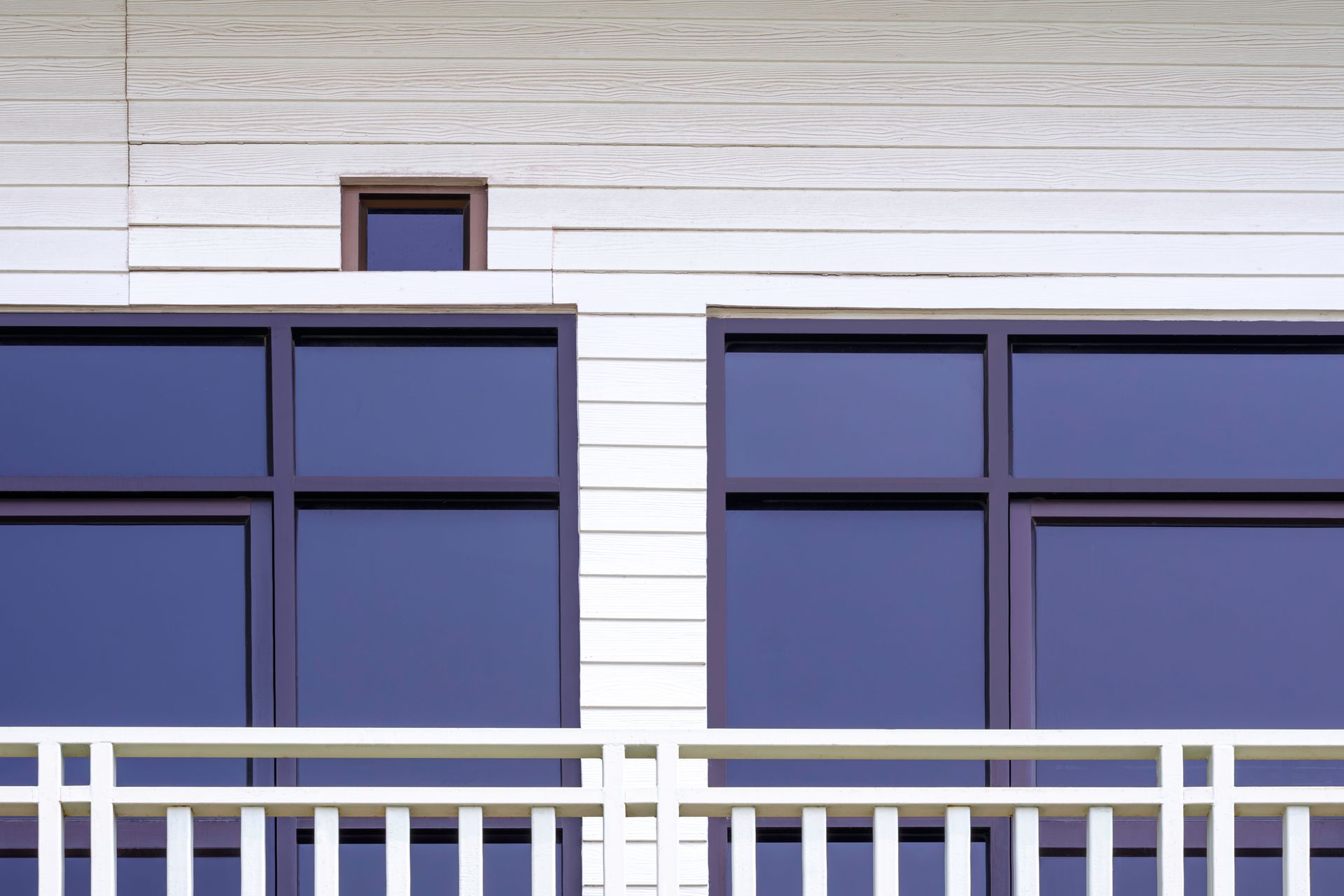 A modern building facade featuring horizontal white siding, two large dark-tinted windows, and a white balcony railing.