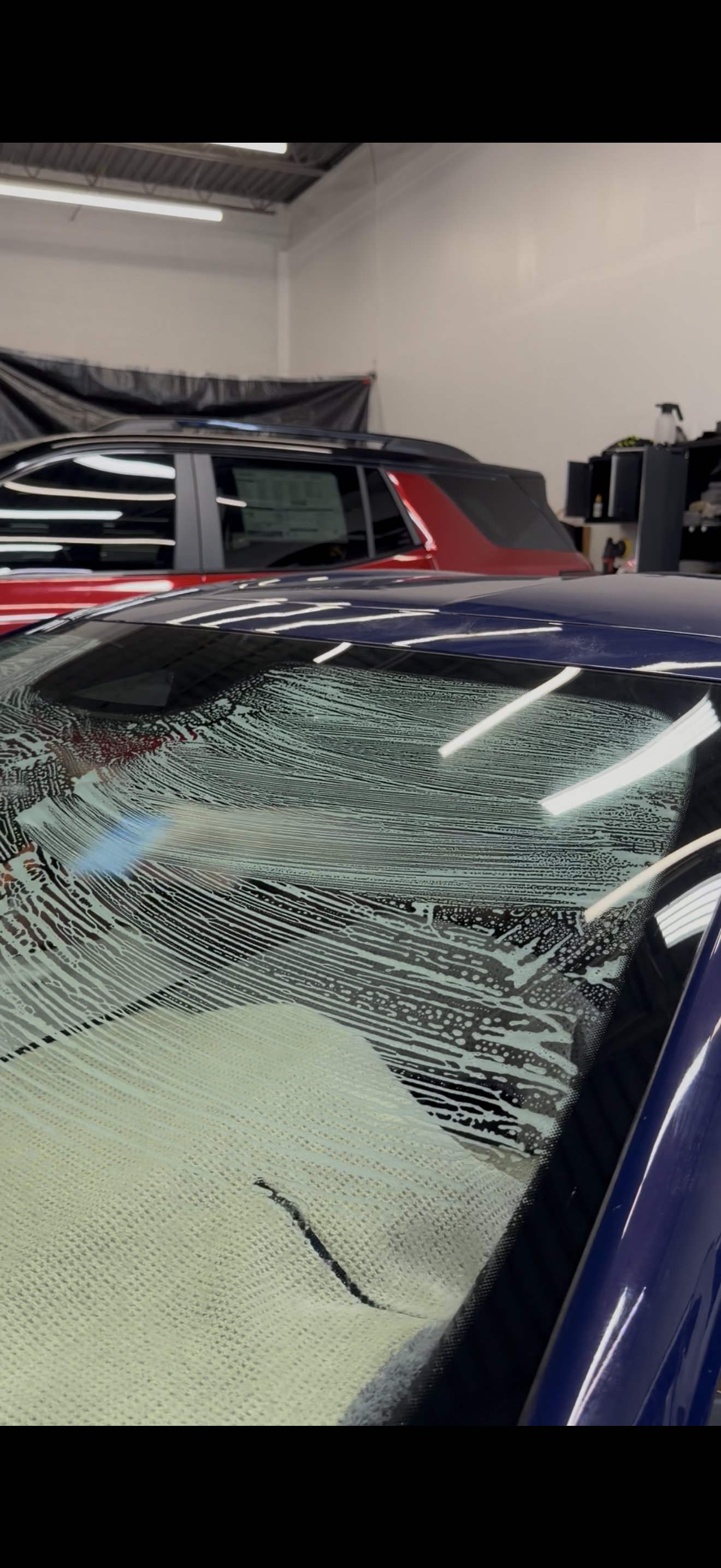 A severely shattered blue car windshield inside a garage with another vehicle visible in the background.