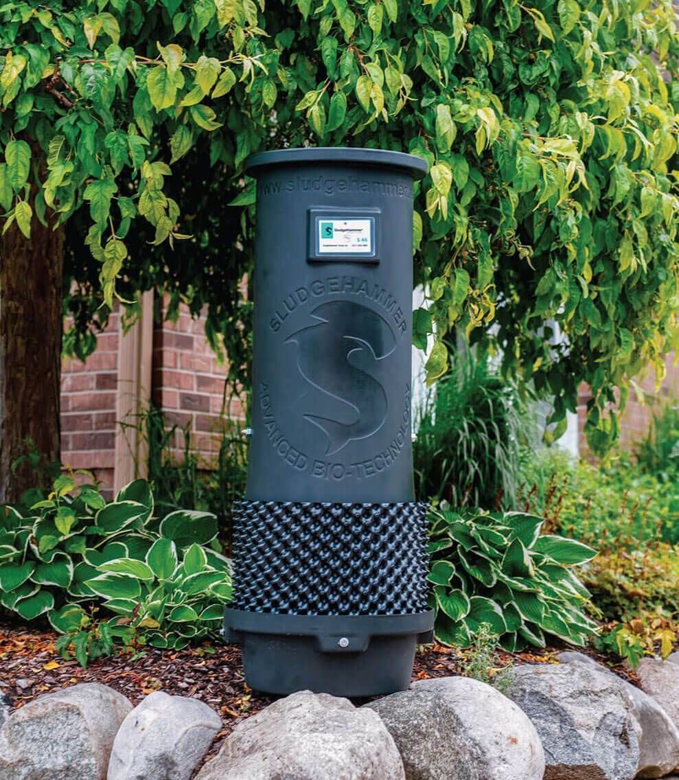 Black outdoor trash bin beside rocks and greenery in a garden setting