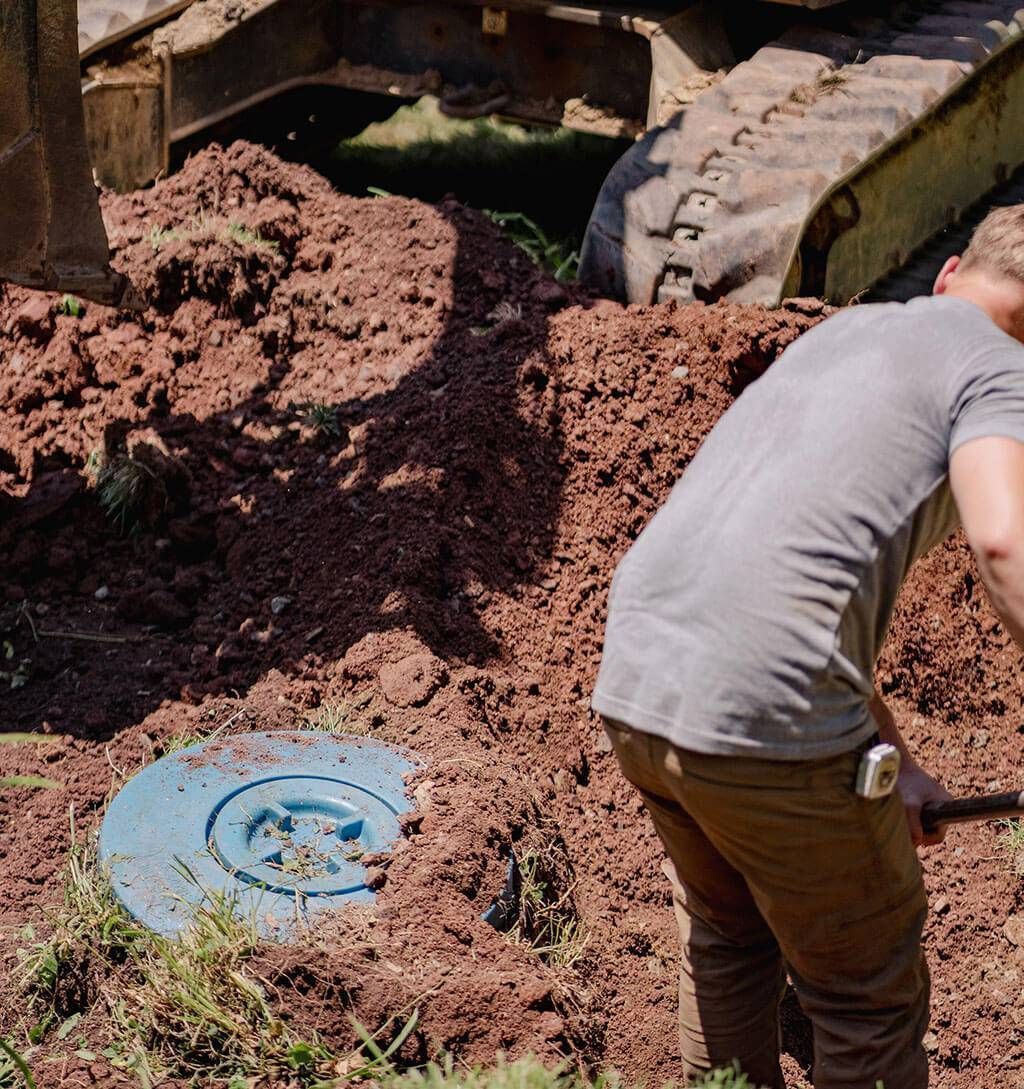 Person working beside a large excavator on a muddy construction site near a blue manhole cover