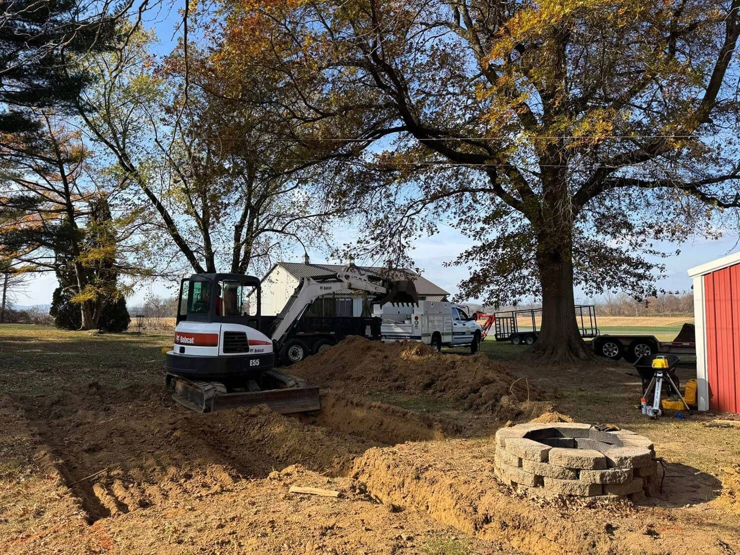 White truck and excavator in a dirt yard under large trees, with a stone fire pit in front.