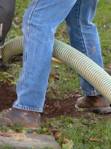 Person in blue jeans and work boots standing over a green corrugated hose on muddy grass.