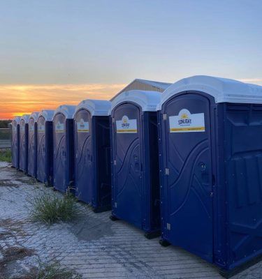 Row of blue portable toilets at sunset beside a paved path and open field