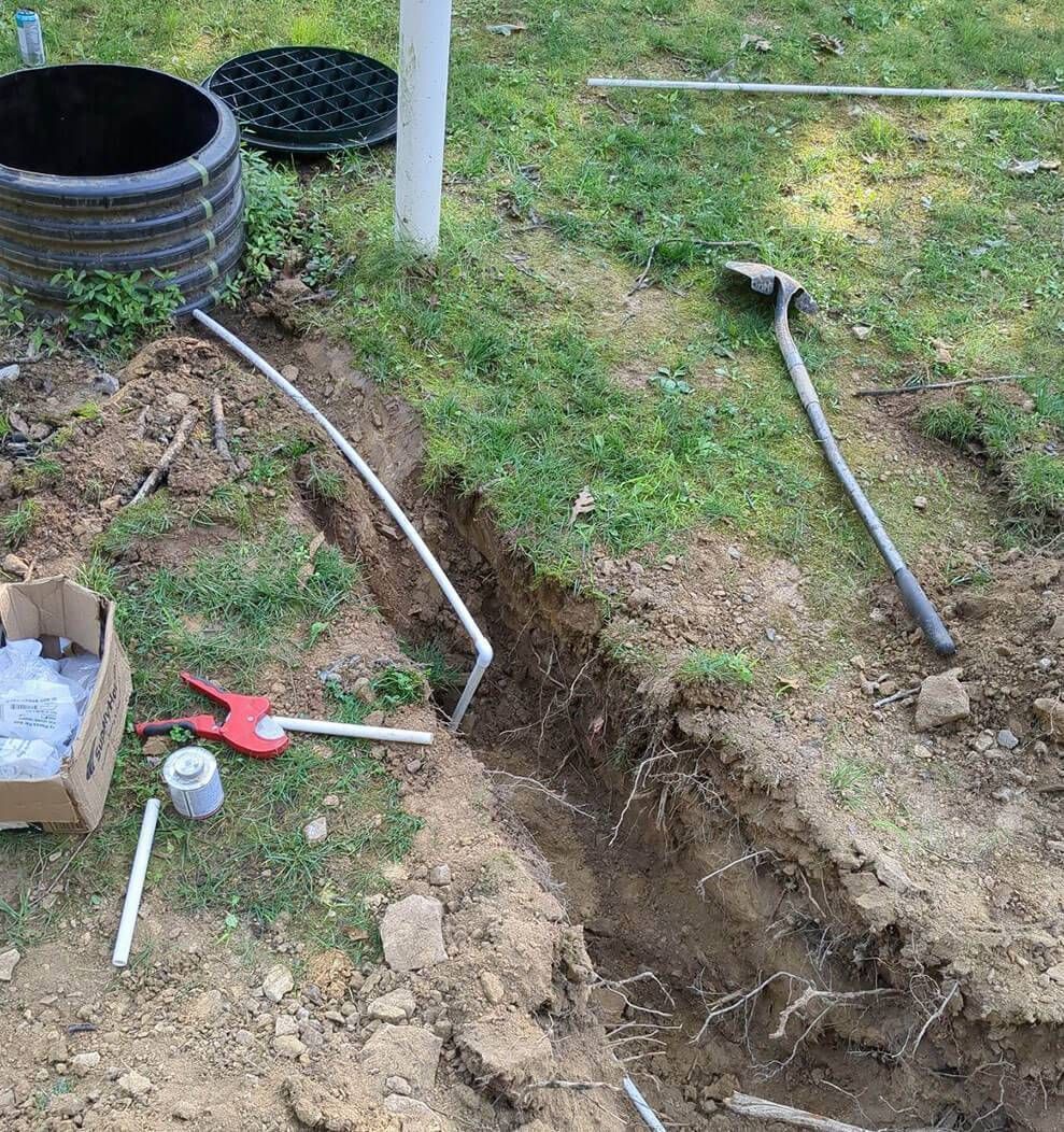 Backyard trench with exposed pipe, hose, and tools beside a black drainage well cover