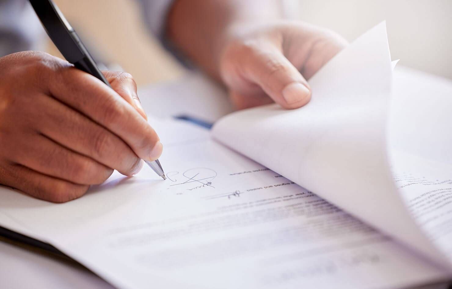 Hands signing a document on a desk with a pen, with pages being turned over