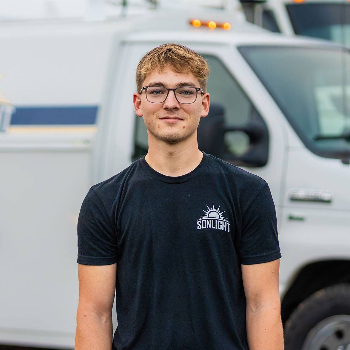 Young man in glasses wearing a black T-shirt stands in front of a white van outdoors