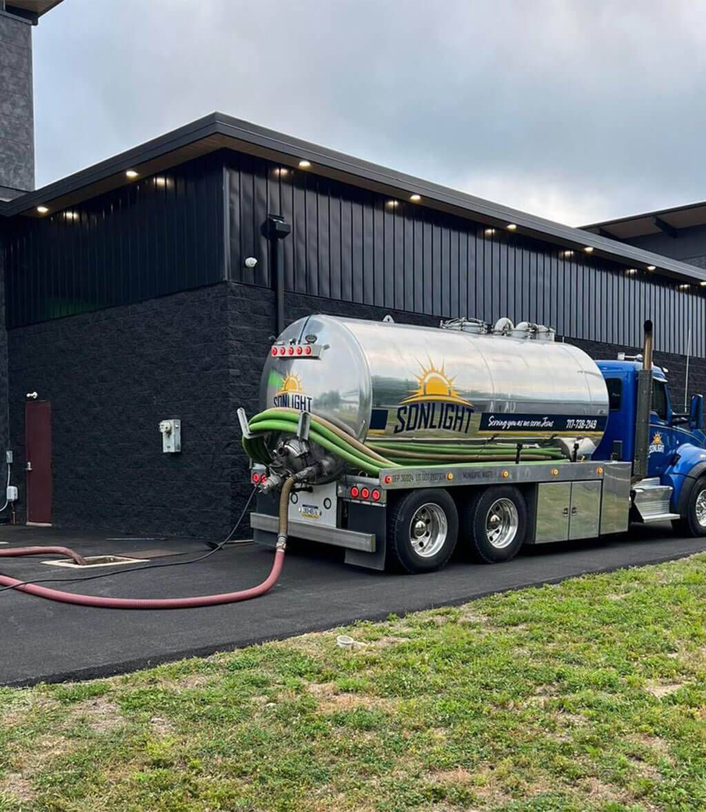Polished silver tanker truck parked beside a black building with hoses connected and grass in the foreground