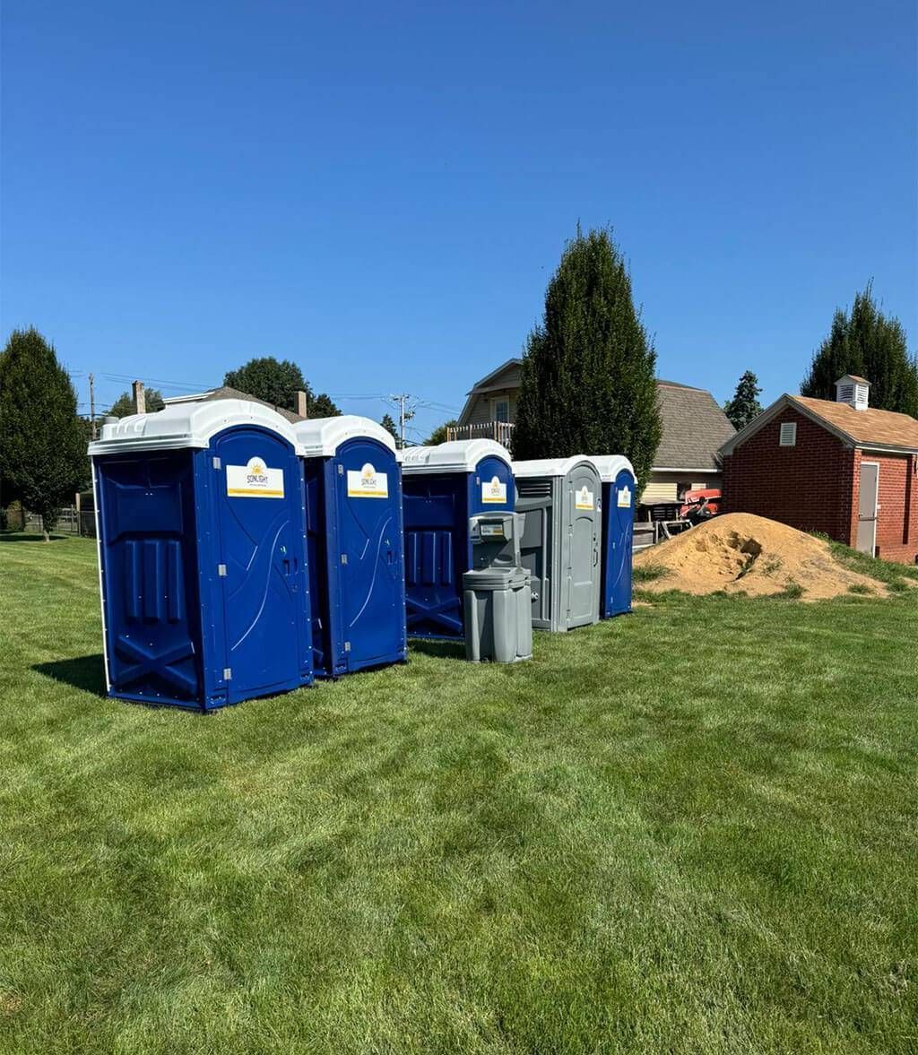 Blue portable toilets and trash bins on a grassy field near a brick building under a clear blue sky