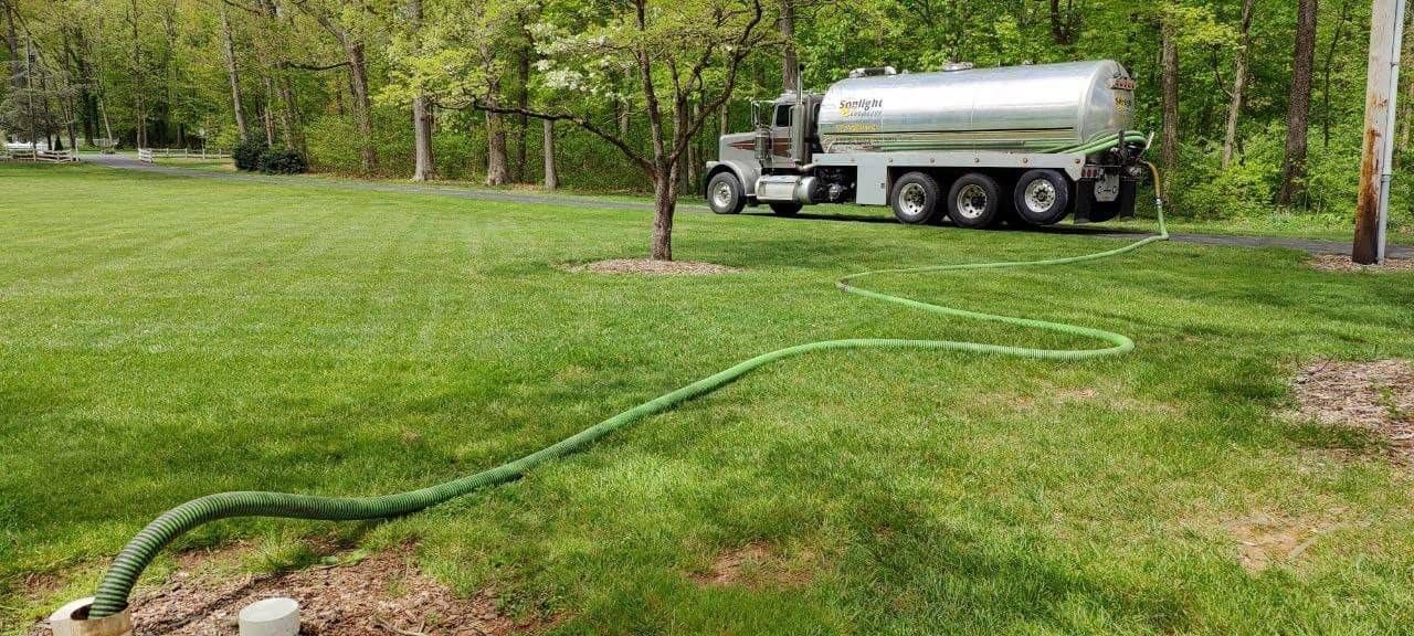 White tanker truck parked on a grassy park lawn beside a hose, with trees in the background