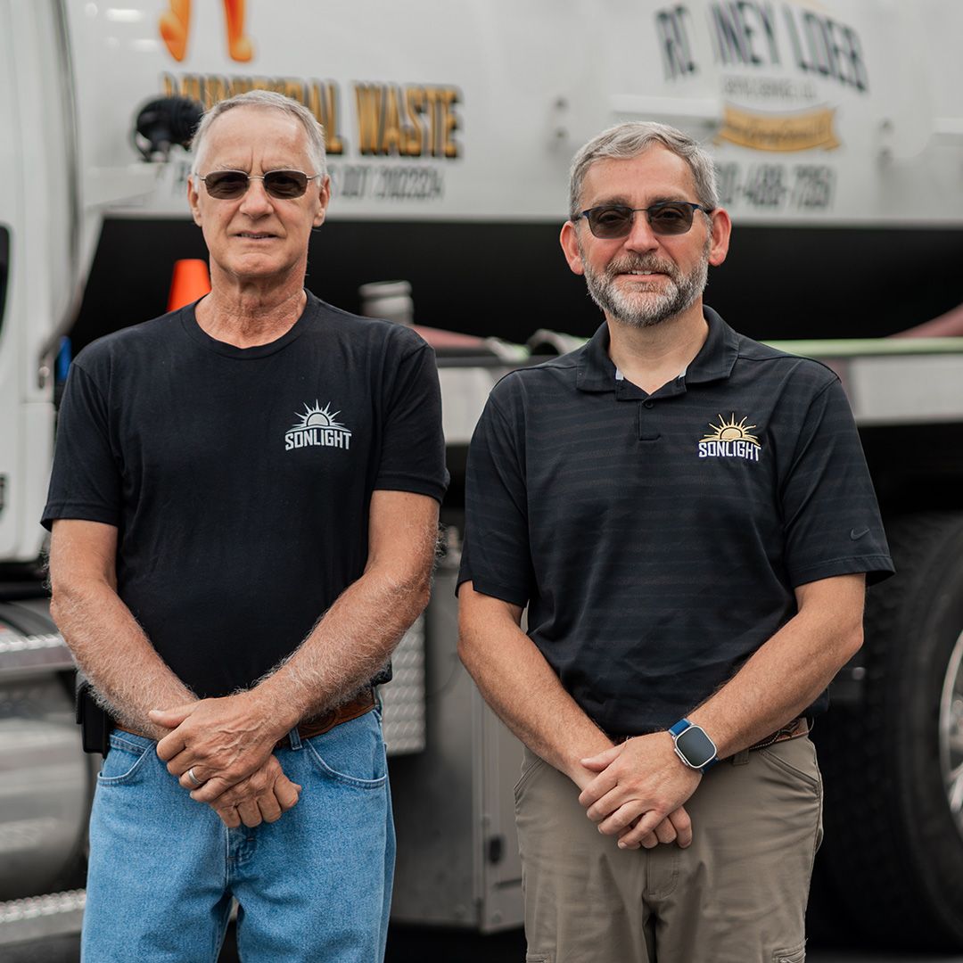 Two men stand in front of a truck, wearing dark shirts and sunglasses, with hands clasped.