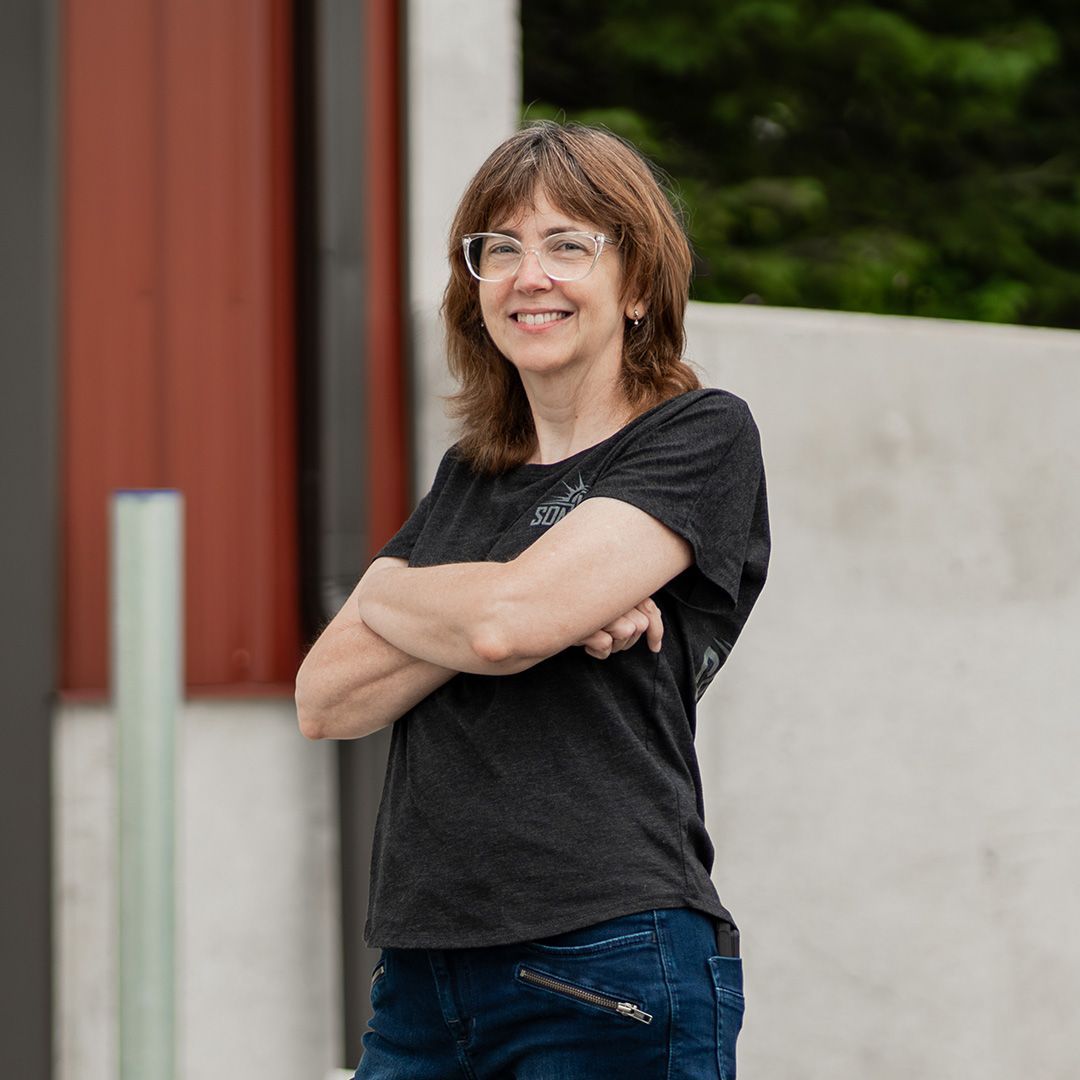 Heather standing outdoors with arms crossed, wearing glasses, a dark shirt, and jeans.