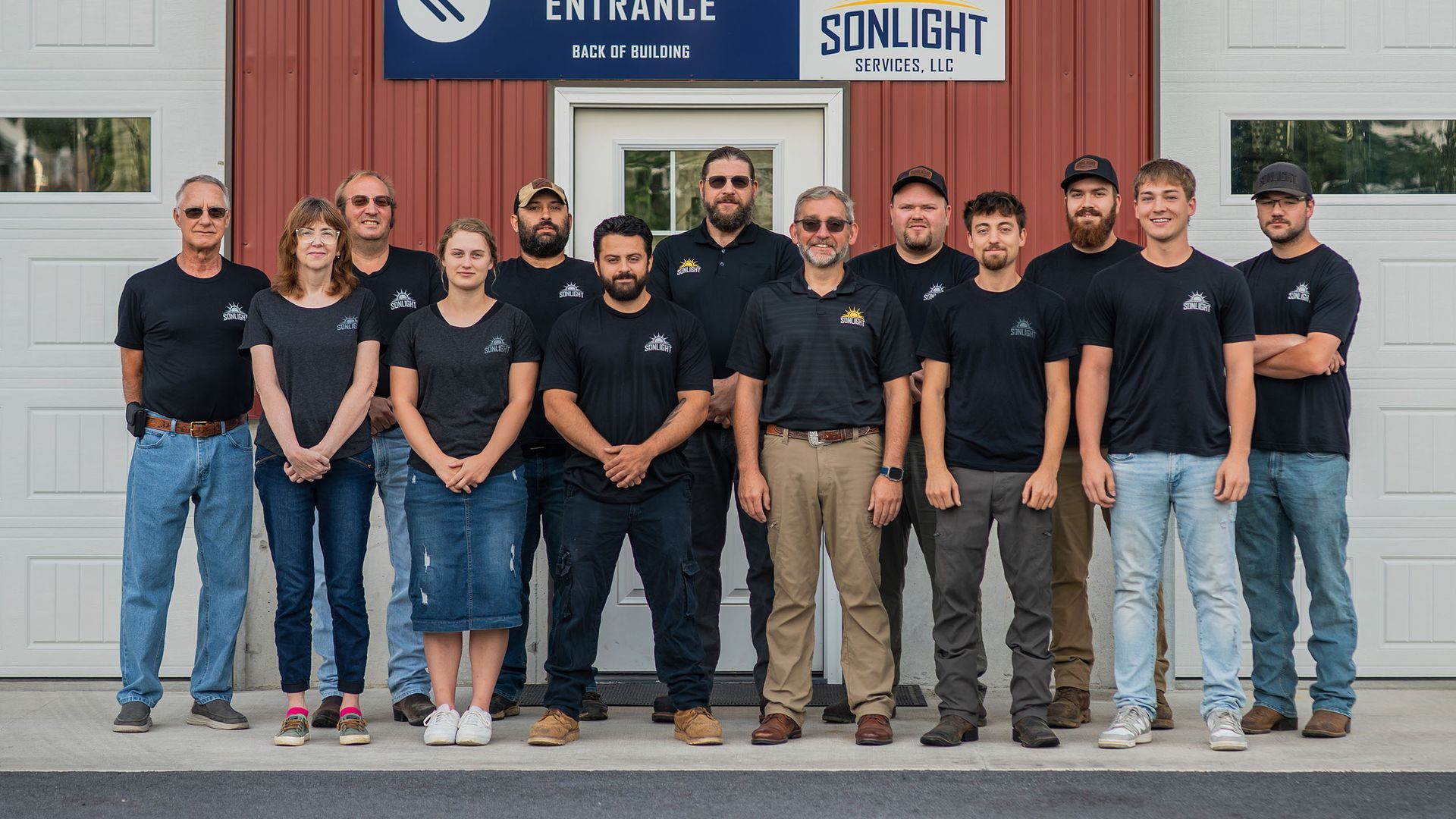 Group of eight people standing outside a red building with a “SUNLIGHT” sign, wearing dark shirts and smiling