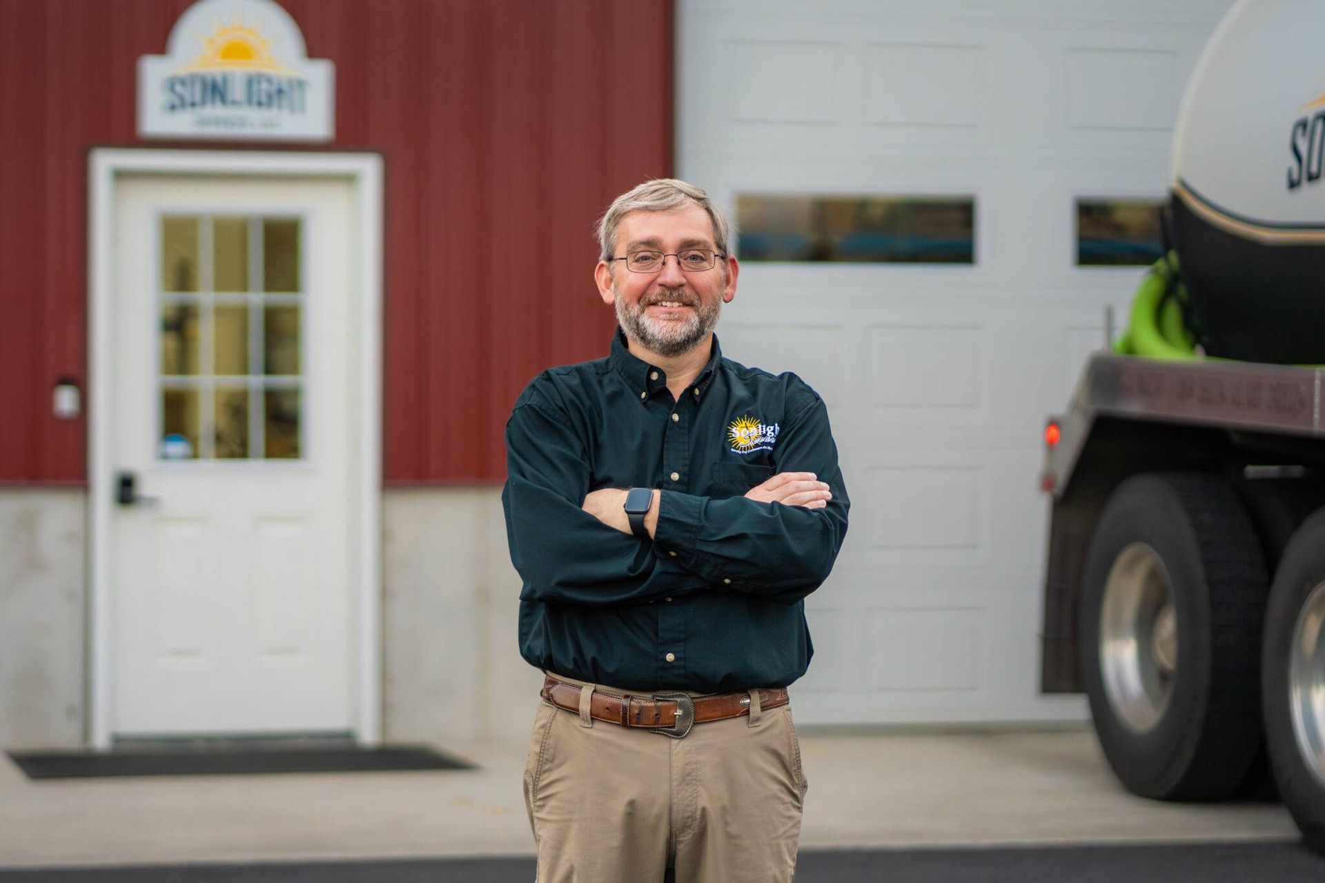 Smiling man with crossed arms stands outside a building beside a truck trailer.