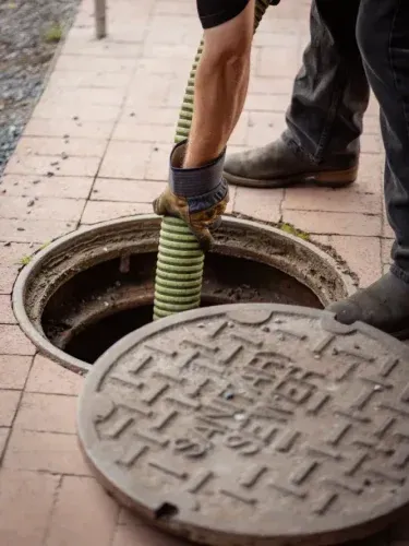 Worker using a green flexible hose to clean a manhole beside an open street cover