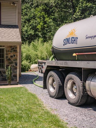 Silver septic truck beside a wooden house on a gravel driveway with green grass and trees nearby