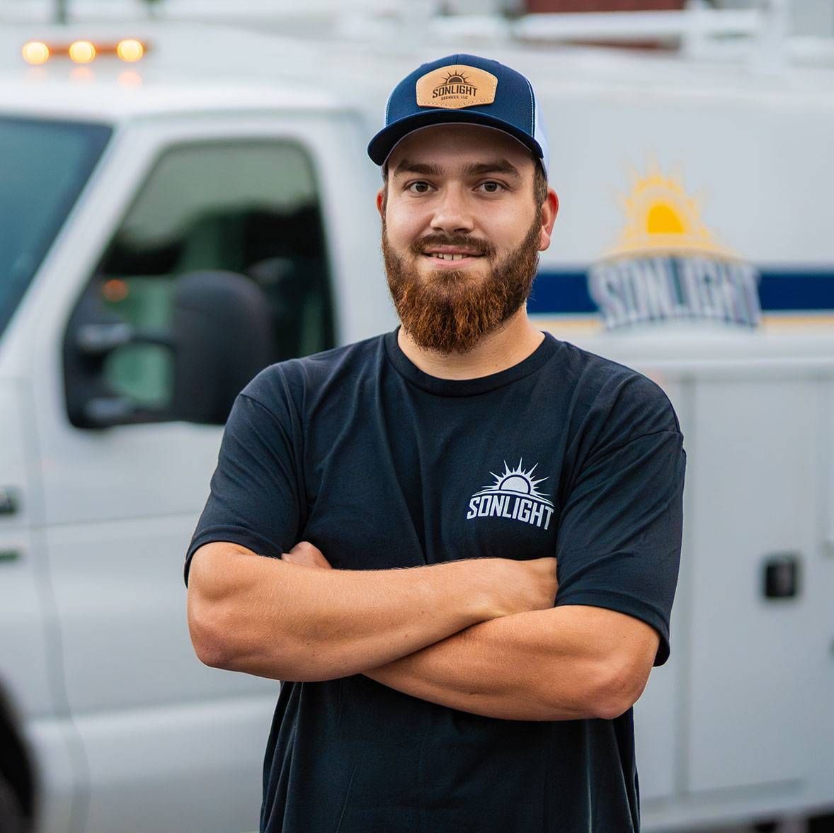 Tyrell in black T-shirt and blue cap standing with arms crossed in front of a white service truck.
