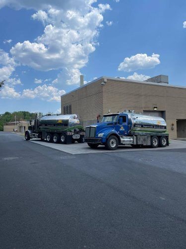 Two tanker trucks parked beside a tan warehouse under a blue sky with clouds.
