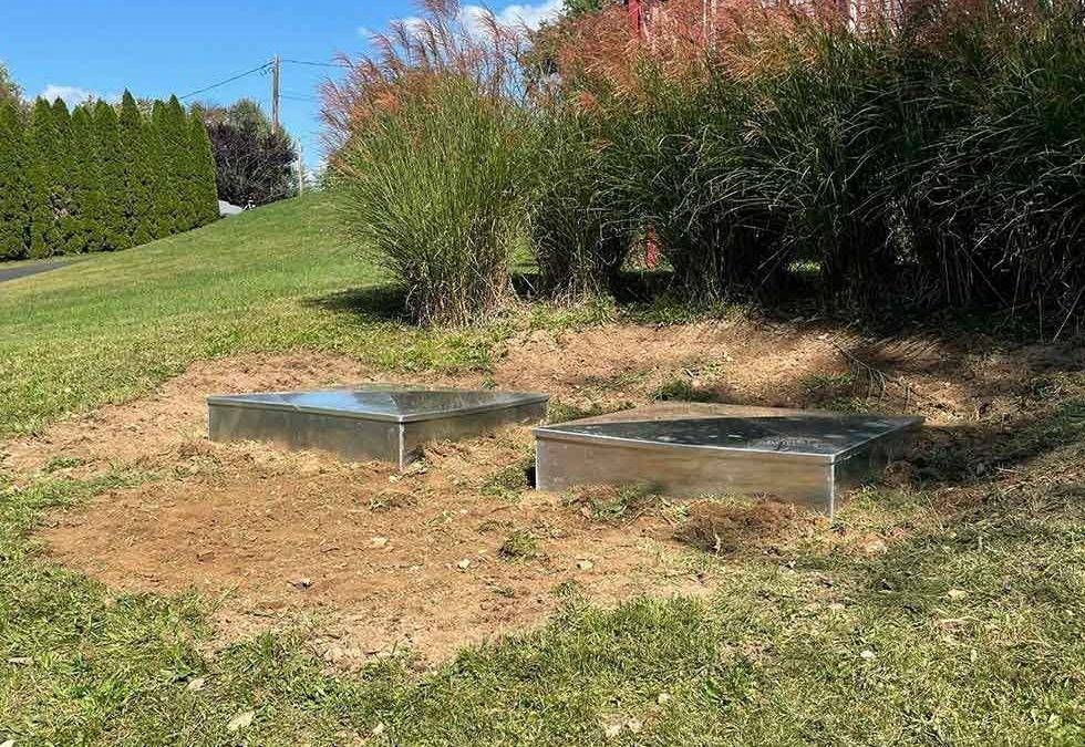 Two low metal boxes on a grassy slope beside shrubs and a hedge under a clear blue sky