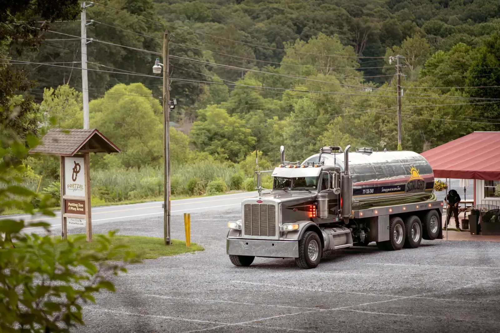 Silver tanker truck parked by a red-roofed building on a gravel lot beside a forested road
