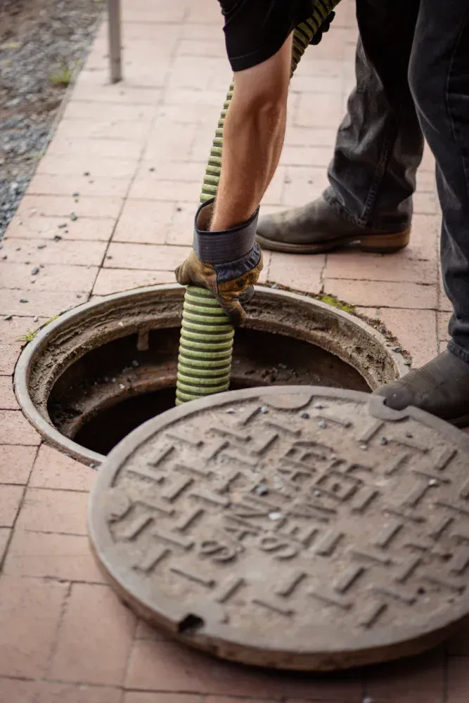 Worker handling green-striped cable at open manhole on sidewalk, with manhole cover nearby