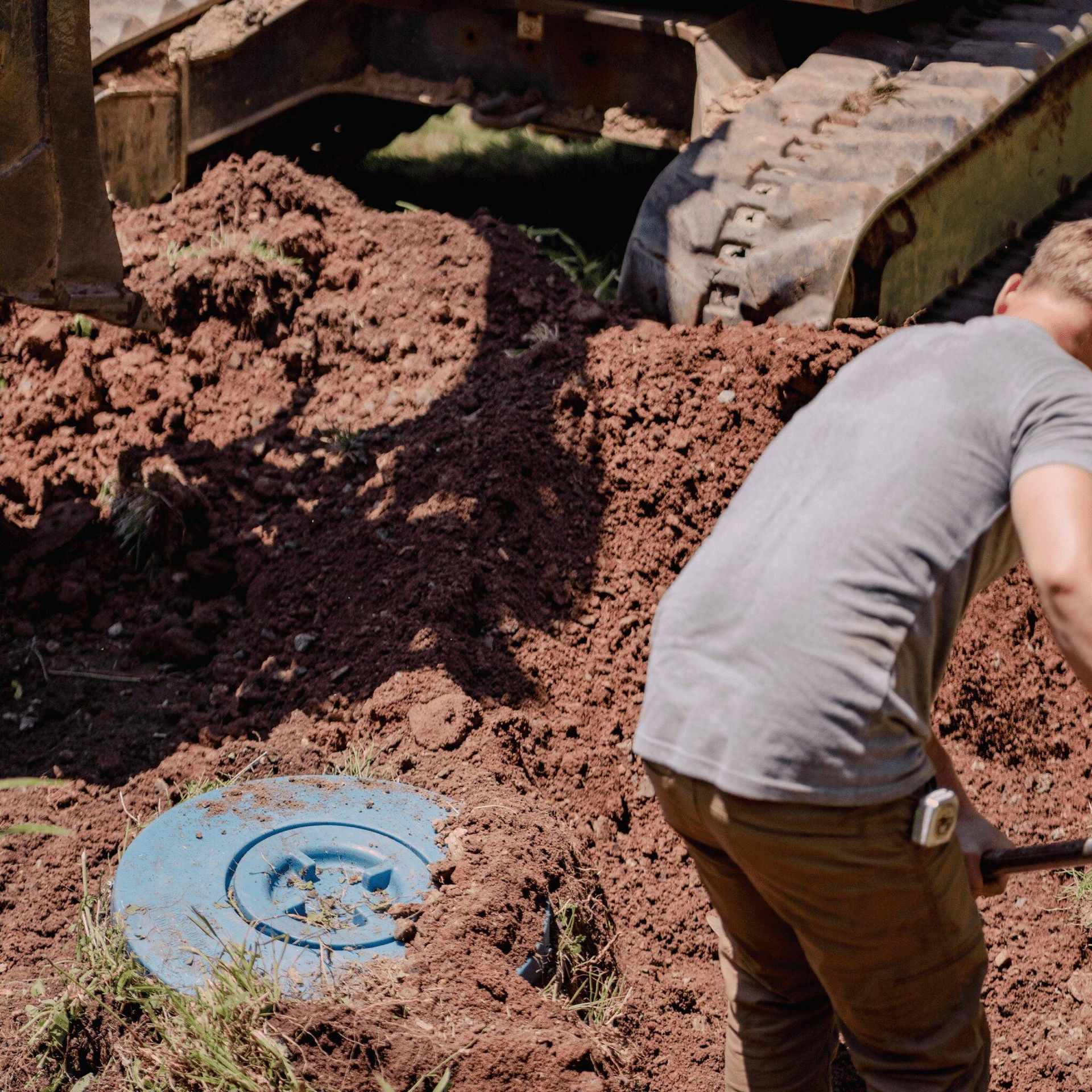 Worker beside excavator digging brown soil near a blue manhole cover