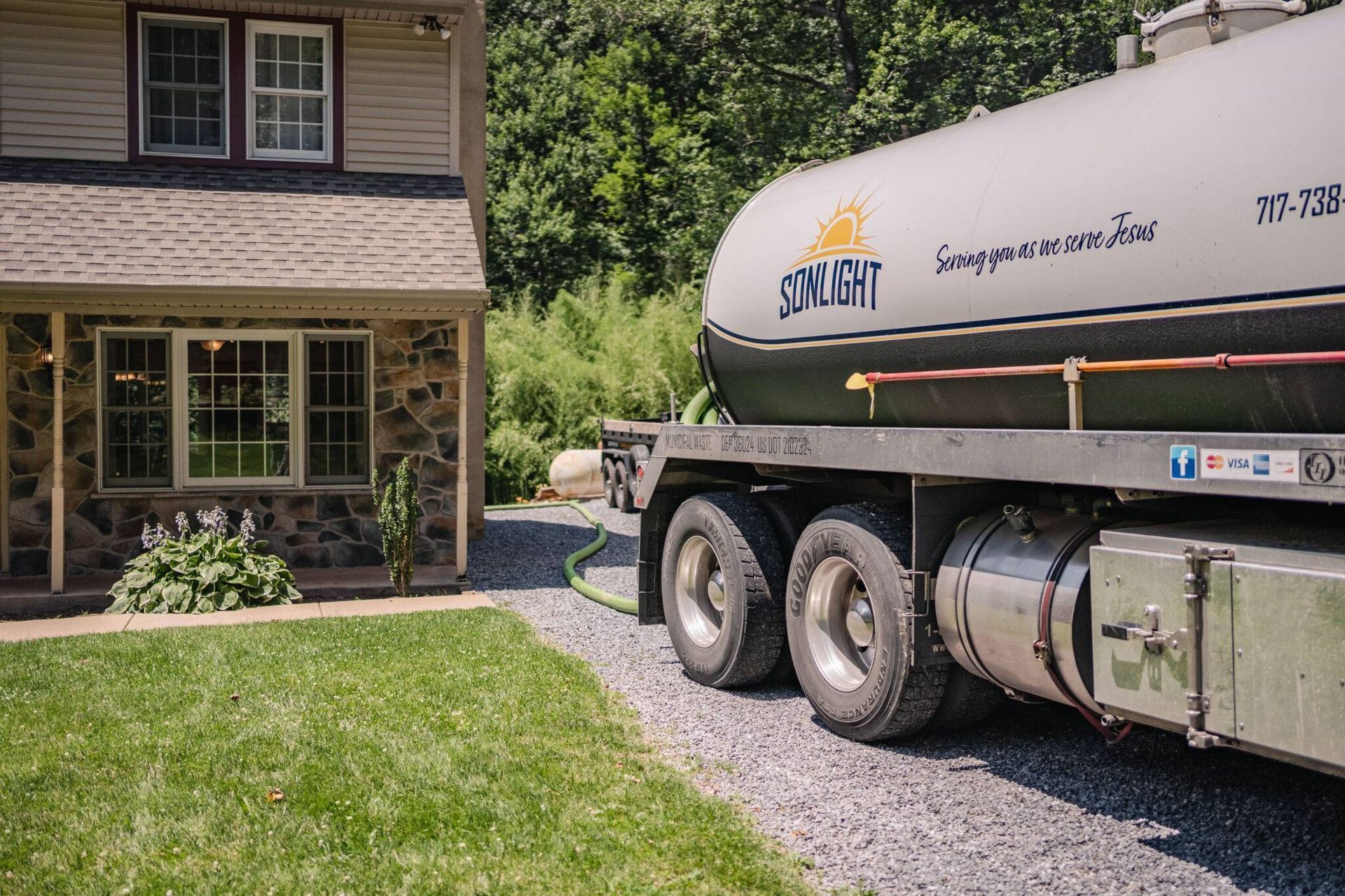 House with large white tanker truck parked on gravel driveway and lawn near woods