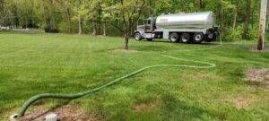 Silver tanker truck parked on a grassy field with a green hose stretched across the lawn