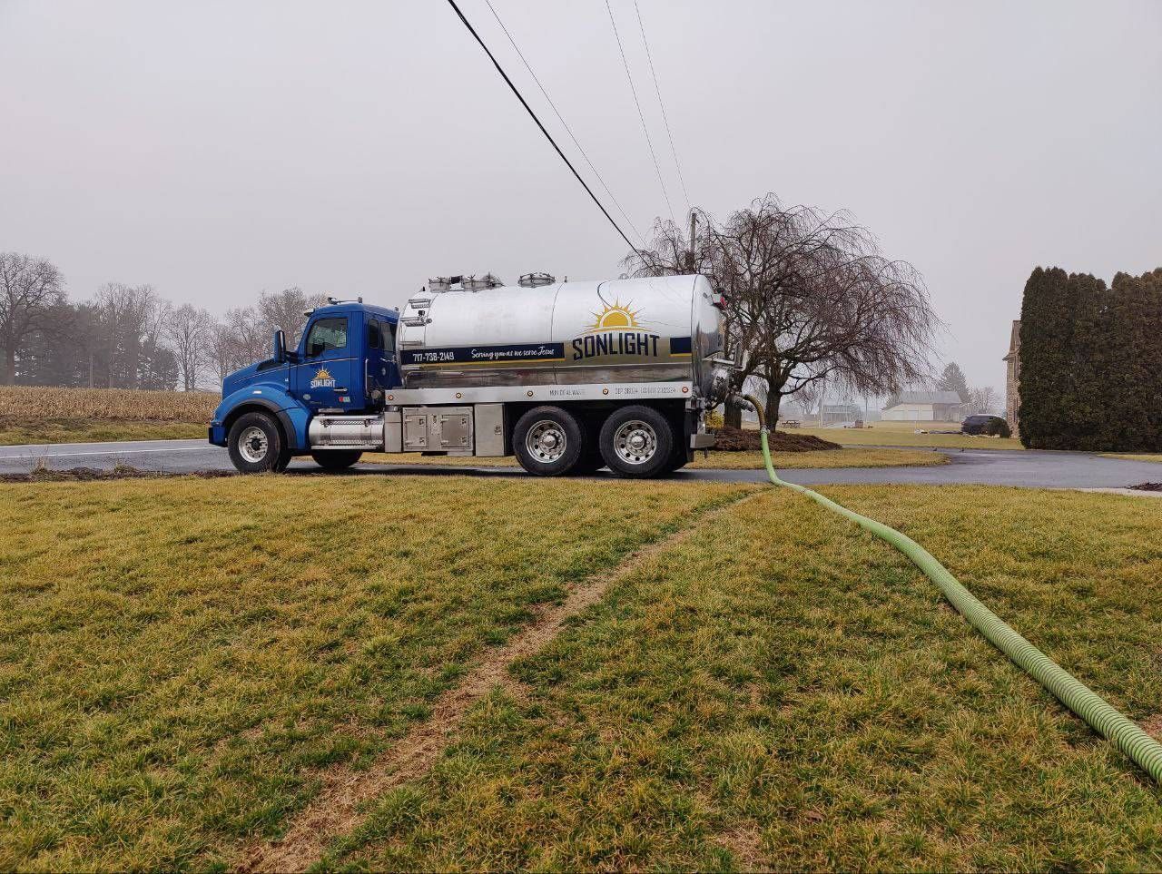 Blue tanker truck on a rainy rural road beside a field, with a hose stretched across the grass.