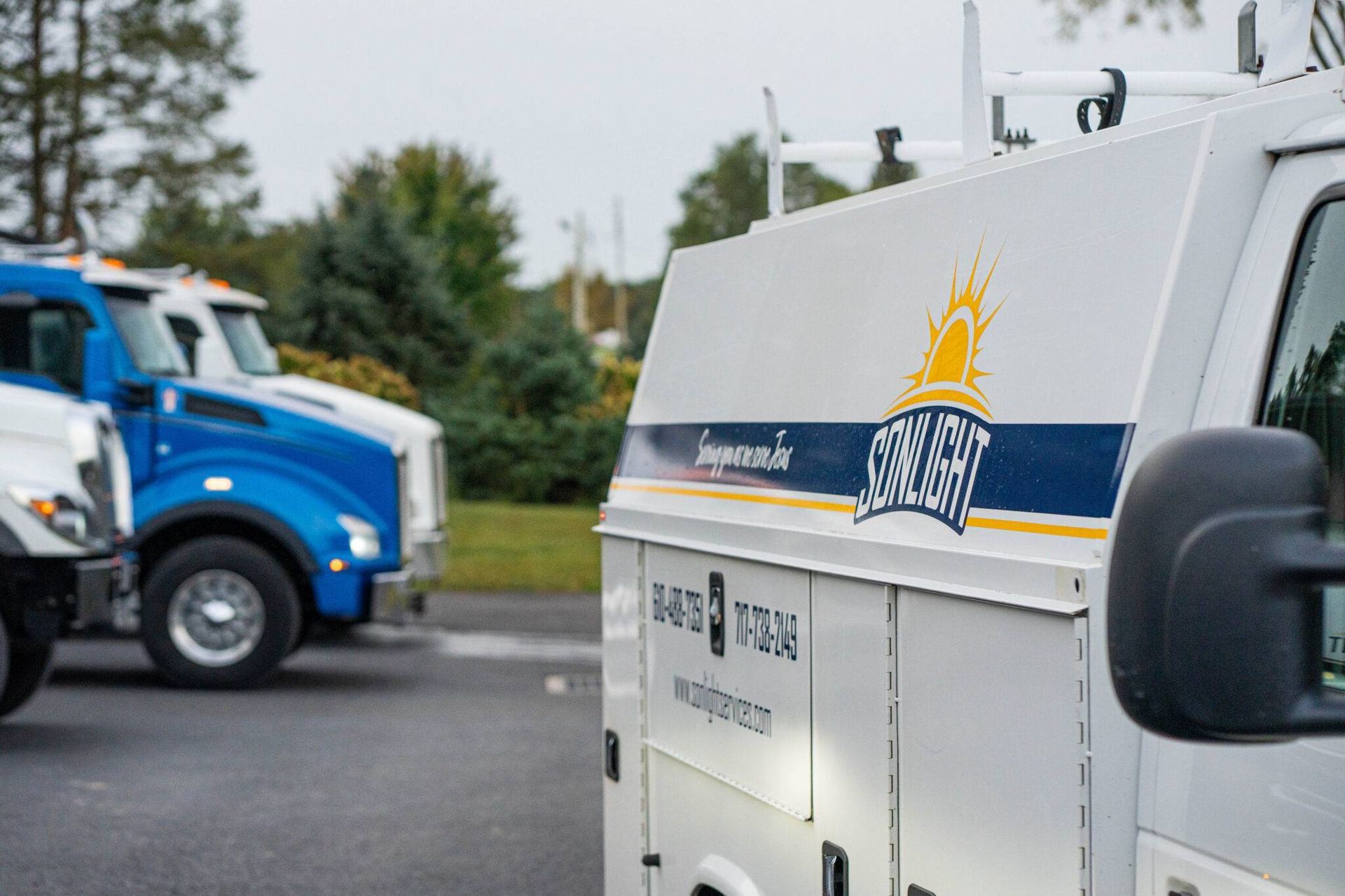 White utility van and blue truck parked outdoors in a lot with overcast sky