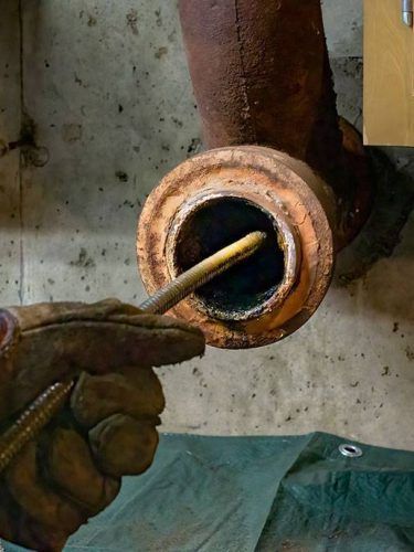 Gloved hand probing a rusted pipe opening with a metal tool against a concrete wall