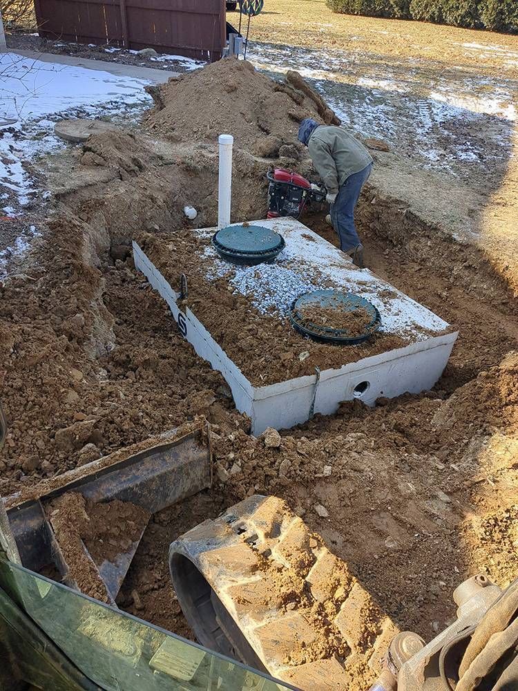 Worker installing septic tank in a backyard excavation, with concrete lids and PVC pipe visible.