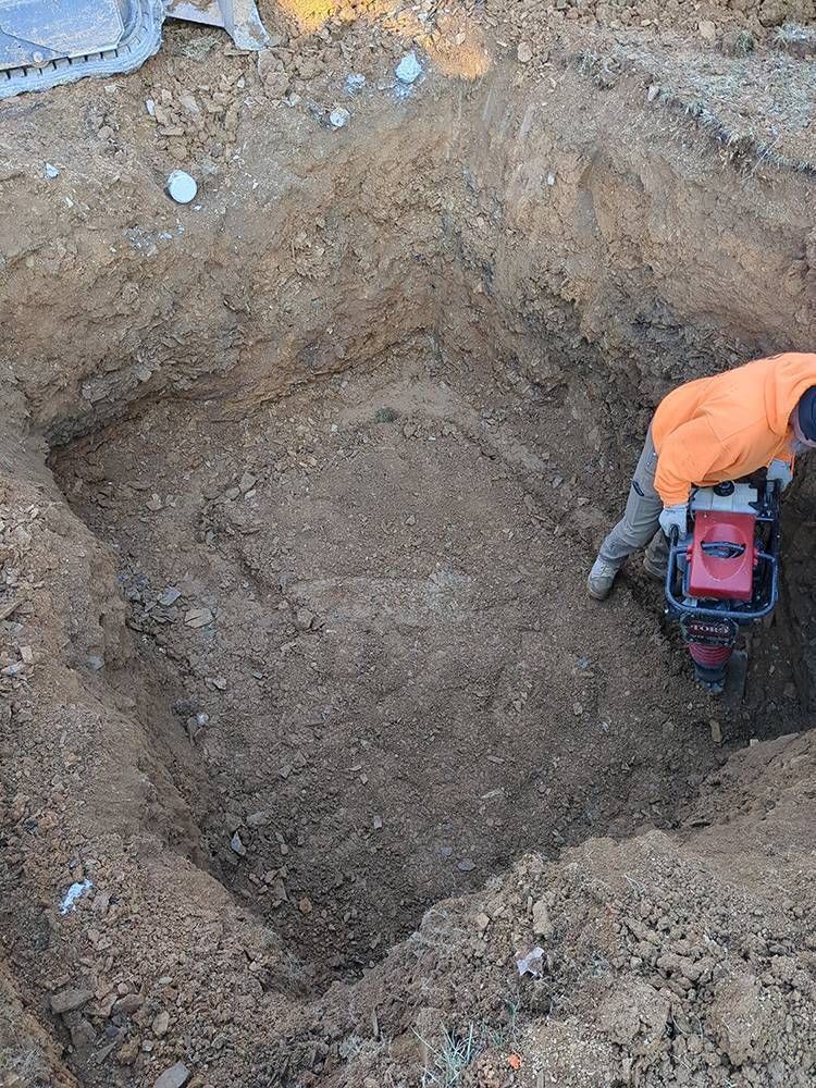 Worker in orange jacket using a machine to dig a large hole in bare ground.